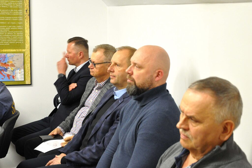 Pięciu mężczyzn siedzi w rzędzie na jasnym tle, ubrani w marynarki i swetry, jeden trzyma kartkę papieru, obok widoczna mapa i fragment tekstu na ścianie.