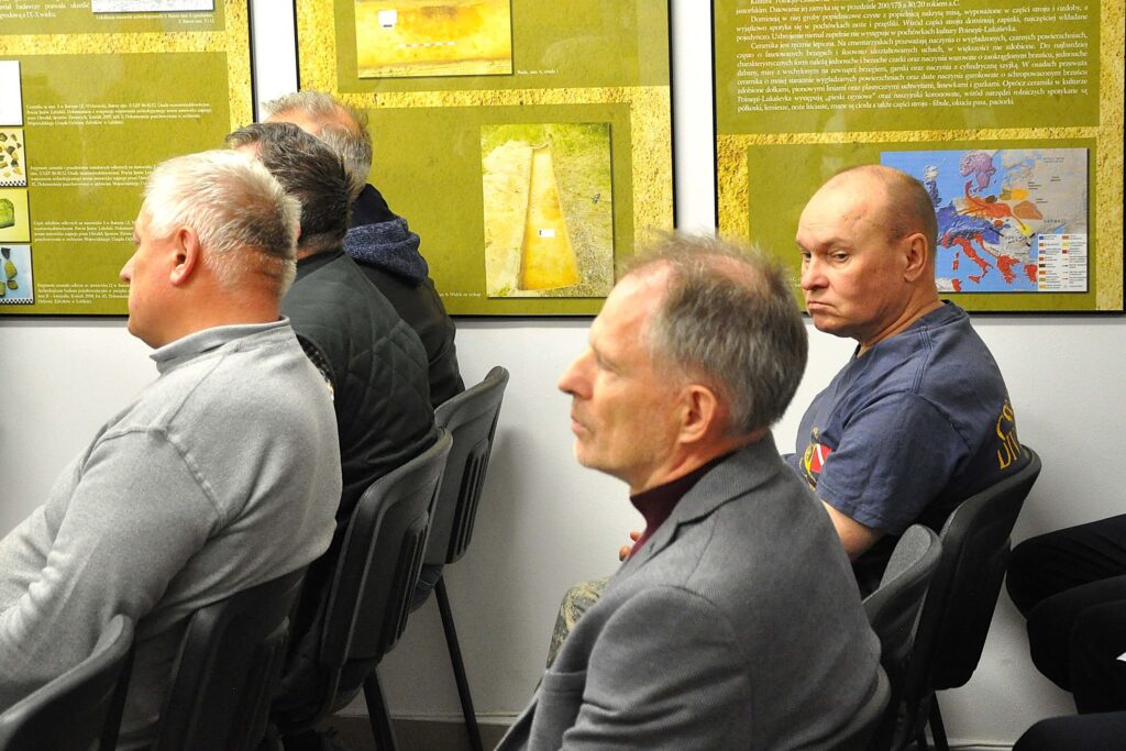 Czterech mężczyzn siedzi na czarnych krzesłach w sali, na ścianie za nimi wiszą żółto-zielone plansze z tekstem i zdjęciami.