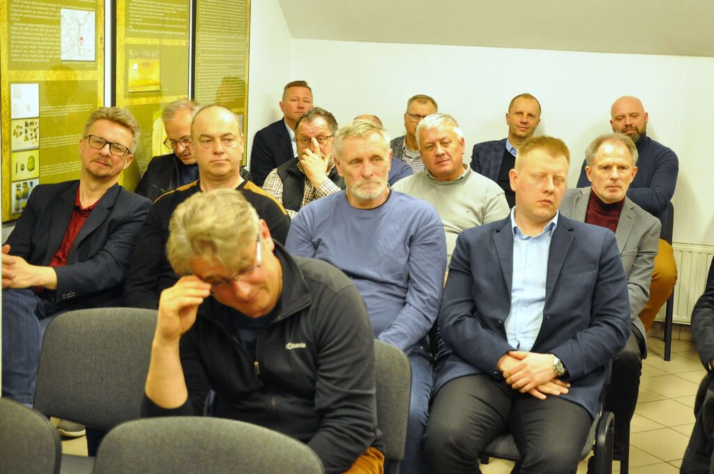 Grupa mężczyzn w różnorodnych ubraniach siedzi na szarych krzesłach w jasnym pomieszczeniu z białymi ścianami i żółtymi planszami na ścianie.