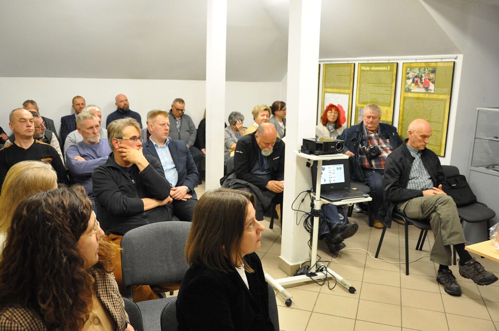 Grupa ludzi siedzących na krzesłach w jasnym pomieszczeniu z białymi ścianami, laptop i projektor na stojaku, tablice z tekstem i zdjęciami na ścianie.