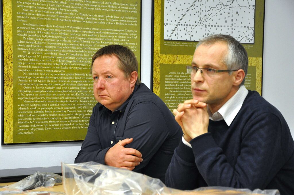 Dwaj mężczyźni siedzący przy stole, za nimi tablice z tekstem i schematami na żółtym tle, jeden w ciemnej koszuli, drugi w granatowym swetrze i okularach.