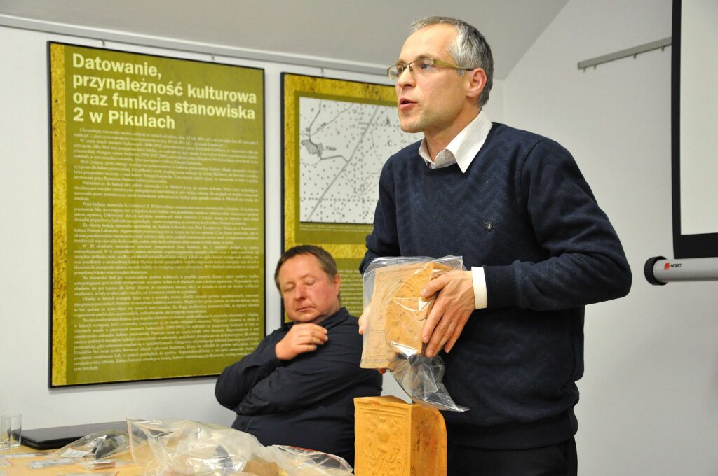 Mężczyzna w granatowym swetrze trzymający glinianą płytkę z ornamentem, drugi mężczyzna siedzi z zamkniętymi oczami, w tle zielono-żółte plansze z tekstem i mapą.