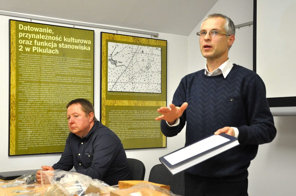 Dwóch mężczyzn w pomieszczeniu z tablicami informacyjnymi na ścianie, jeden siedzi, drugi stoi i gestykuluje trzymając segregator.