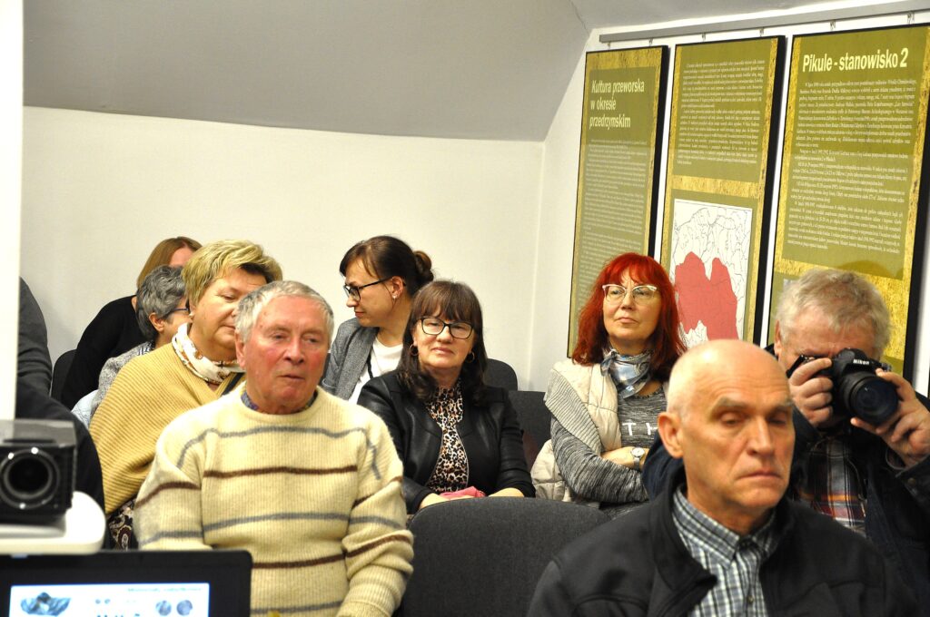 Grupa ludzi siedzących na krzesłach w pomieszczeniu z białą ścianą i tablicami informacyjnymi na żółtym tle. Jeden mężczyzna fotografuje aparatem.