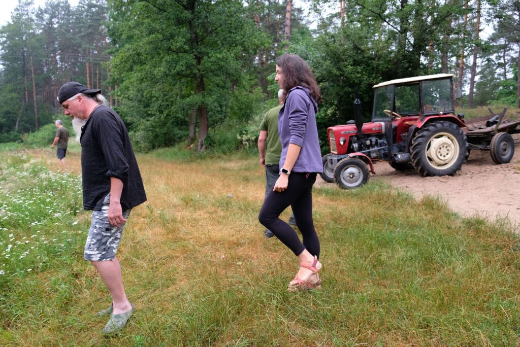 Cztery osoby na trawie i polu kwiatów, w tle zielone drzewa, czerwony traktor z przyczepą na ziemi.