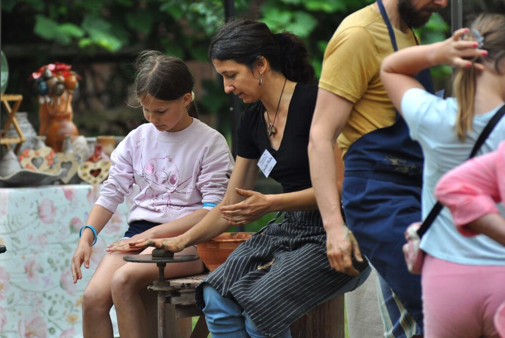 Kobieta i dziewczynka formują glinę na kole garncarskim, obok mężczyzna w musztardowej koszulce i dziewczynka z jasnym włosem, w tle zielone liście i ceramiczne ozdoby.