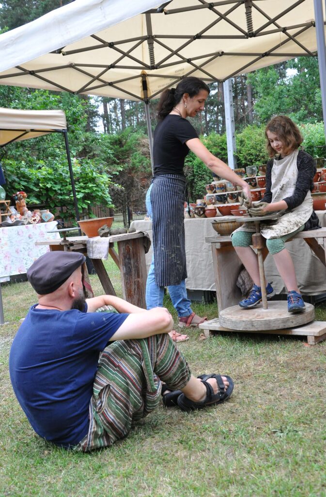 Kobieta i dziecko przy kole garncarskim pod białym namiotem, mężczyzna siedzi na trawie, w tle stół z ceramicznymi naczyniami, zielone drzewa i krzewy.