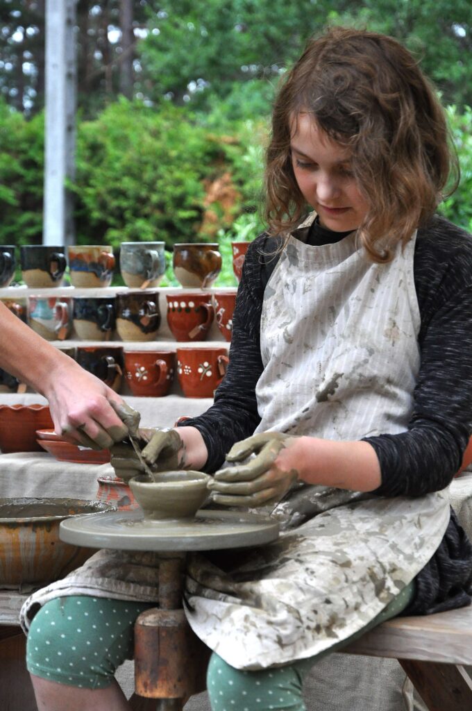 Dziewczynka z brązowymi, falującymi włosami w zielonych spodniach w białej, brudnej od gliny fartuchu formuje gliniany kubek na kole garncarskim, w tle ceramiczne kubki.