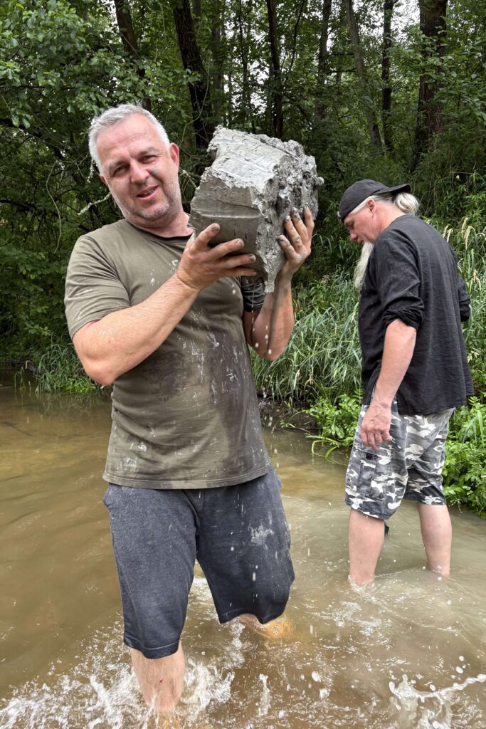 Dwaj mężczyźni stojący w wodzie przy brzegu, jeden trzyma duży kawałek mokrego betonu, drugi ubrany w czarną bluzę i moro szorty stoi bokiem, w tle gęste zielone drzewa.