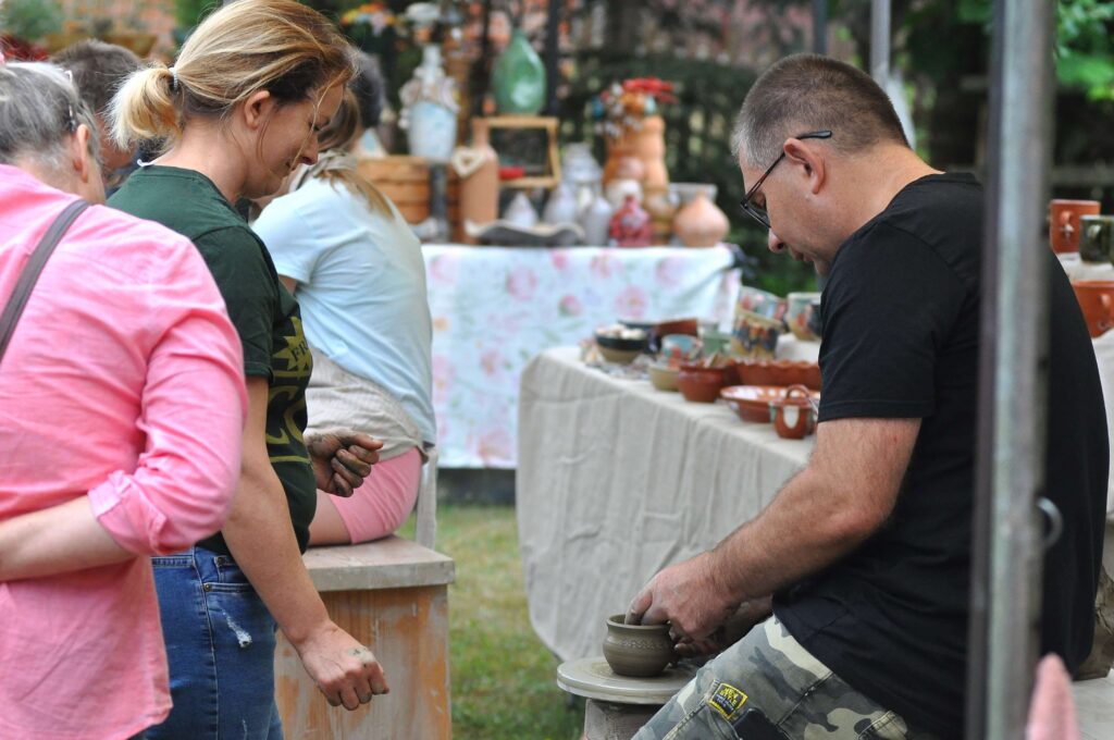 Mężczyzna w czarnej koszulce formujący gliniany dzbanek na kole garncarskim, obok kobiety obserwujące jego pracę, w tle stół z ceramiką i rozmyte rośliny.