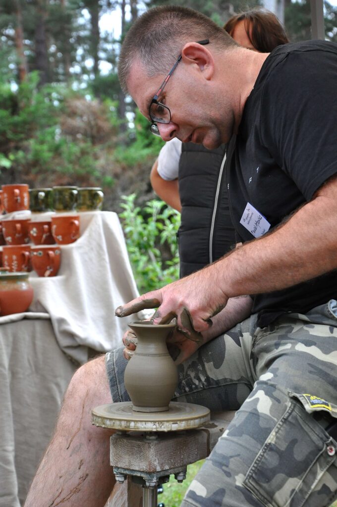 Mężczyzna w okularach i czarnej koszulce formuje gliniany wazon na kole garncarskim, siedząc na ławce, w otoczeniu zieleni i stołu z ceramicznymi kubkami.