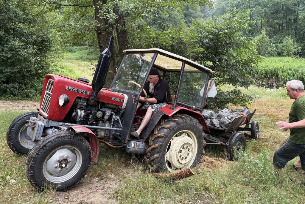 Czerwony traktor z przyczepą pełną drewna, mężczyzna w czarnej koszuli siedzi za kierownicą, drugi mężczyzna w zielonej koszulce stoi obok, zielone drzewa i trawa w tle.
