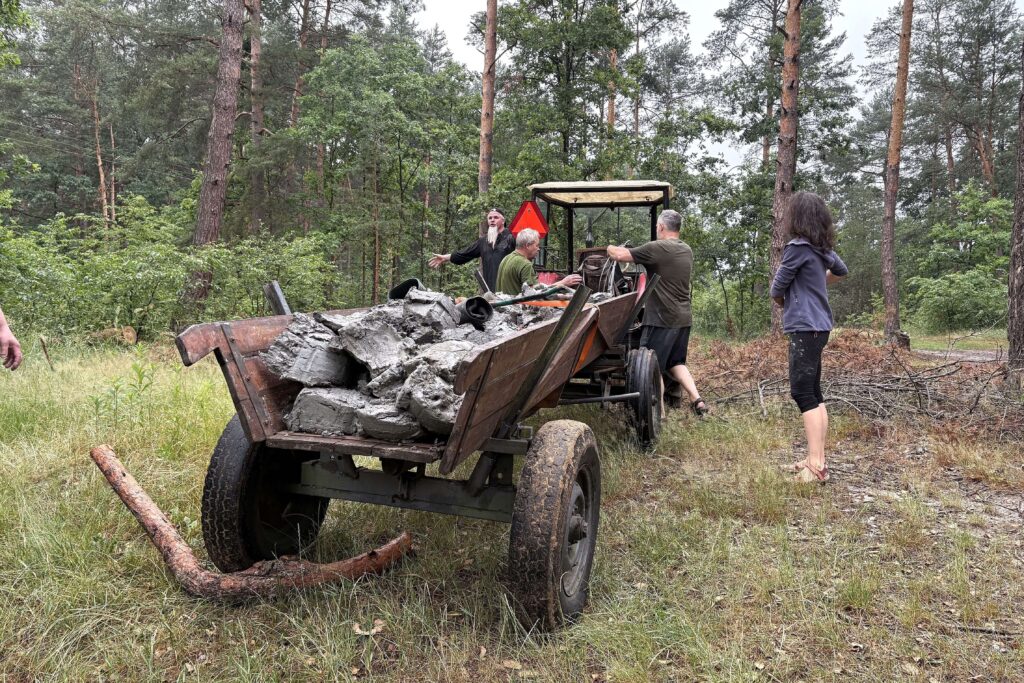 Trzy osoby przy starej przyczepie ciągniętej przez traktor pełnej dużych brył betonu, las sosnowy w tle, kobieta w bluzie i krótkich spodniach stoi obok.