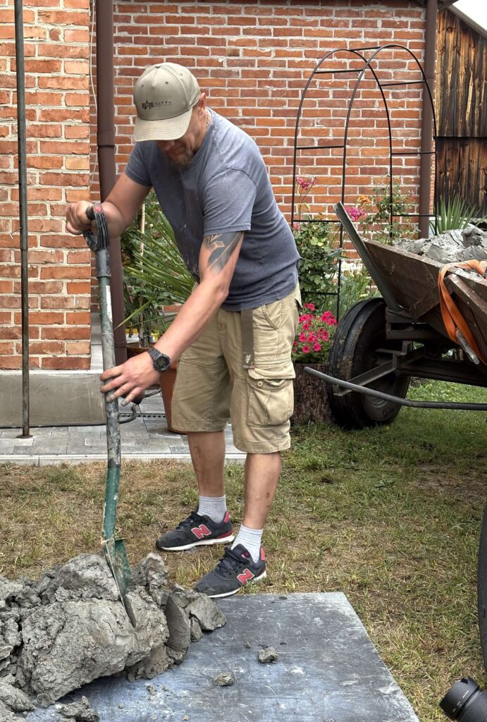 Mężczyzna w czapce, szarej koszulce i beżowych szortach miesza beton zieloną łopatą na trawie, obok taczka z betonem, w tle ceglany mur i kwiaty.