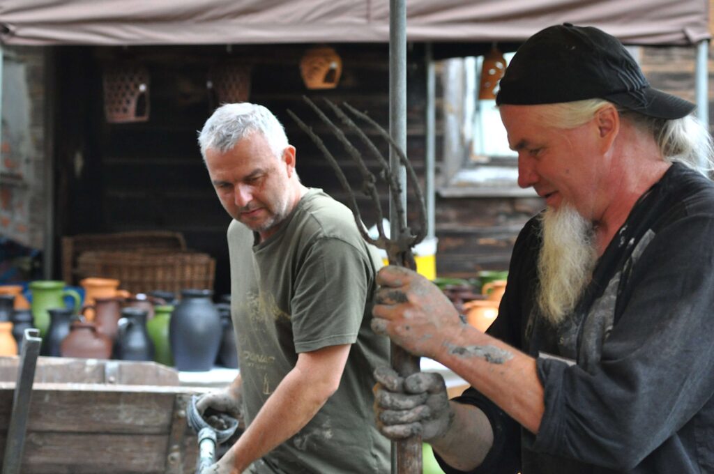 Dwóch mężczyzn z ubrudzionymi rękami, jeden trzyma widły, w tle kolorowe ceramiczne dzbanki i drewniana ściana.