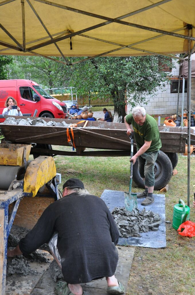 Pod namiotem dwie osoby mieszają i przekładają szarą masę budowlaną, w tle czerwony samochód i ludzie siedzący przy drewnianym stole z glinianymi naczyniami, drzewa, trawa.