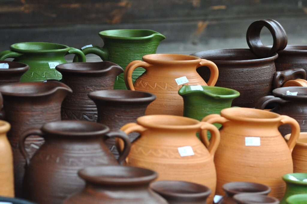 Ceramiczne dzbanki i garnki w kolorach zielonym, brązowym i terakoty z cenami na białych naklejkach, ułożone na stole na tle drewnianej powierzchni.