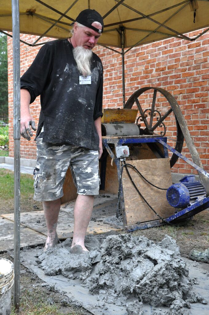 Mężczyzna w czarnej bluzie i kamuflażowych spodniach stoi boso na mokrym, szarym betonie pod żółtym namiotem obok maszyny z dużym kołem.