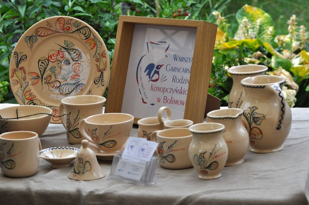 Ceramiczne naczynia w beżowym kolorze z wielobarwnymi, roślinnymi wzorami, ustawione na jasnym obrusie, za nimi zieleń roślin i tabliczka z napisem w drewnianej ramie.