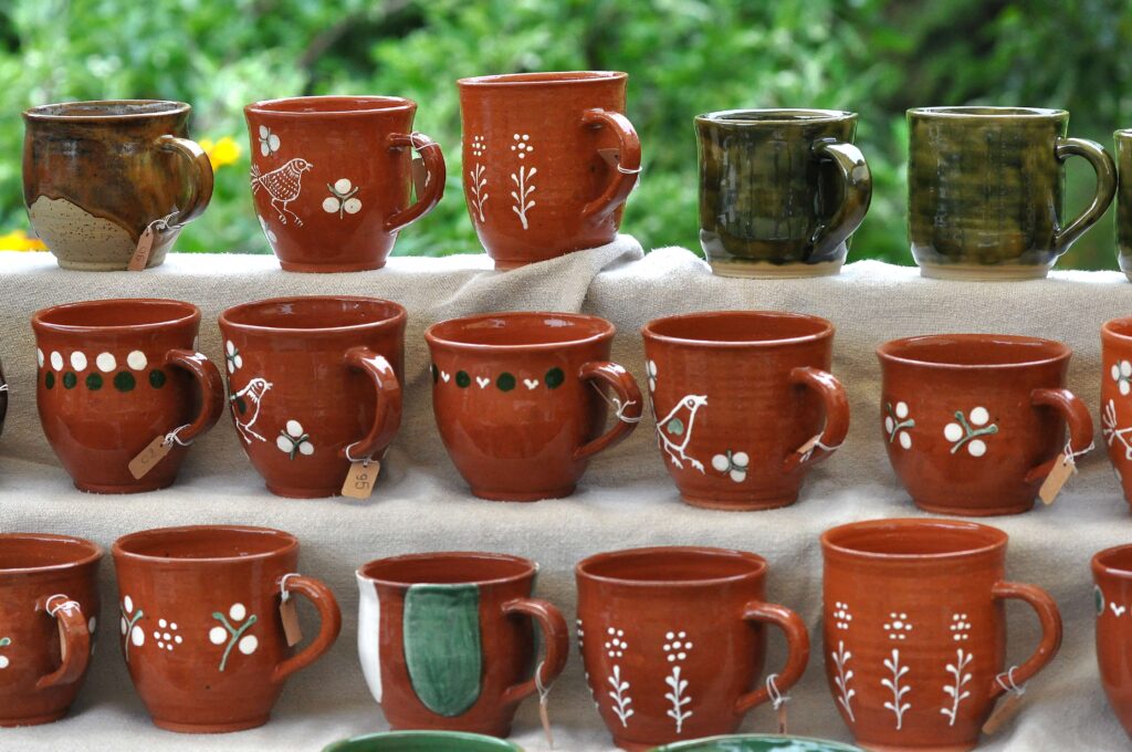 Rzędy ceramicznych kubków w kolorach czerwonym, brązowym i zielonym, niektóre z białymi wzorami roślinnymi i zwierzęcymi, ustawione na jasnej tkaninie na tle rozmytej zieleni.