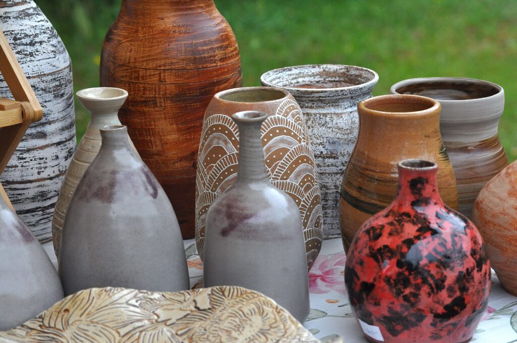 Zestaw naczyń ceramicznych o różnych kształtach i kolorach, m.in. szare, brązowe, czerwono-czarne, z białymi wzorami, na tle zielonej trawy.
