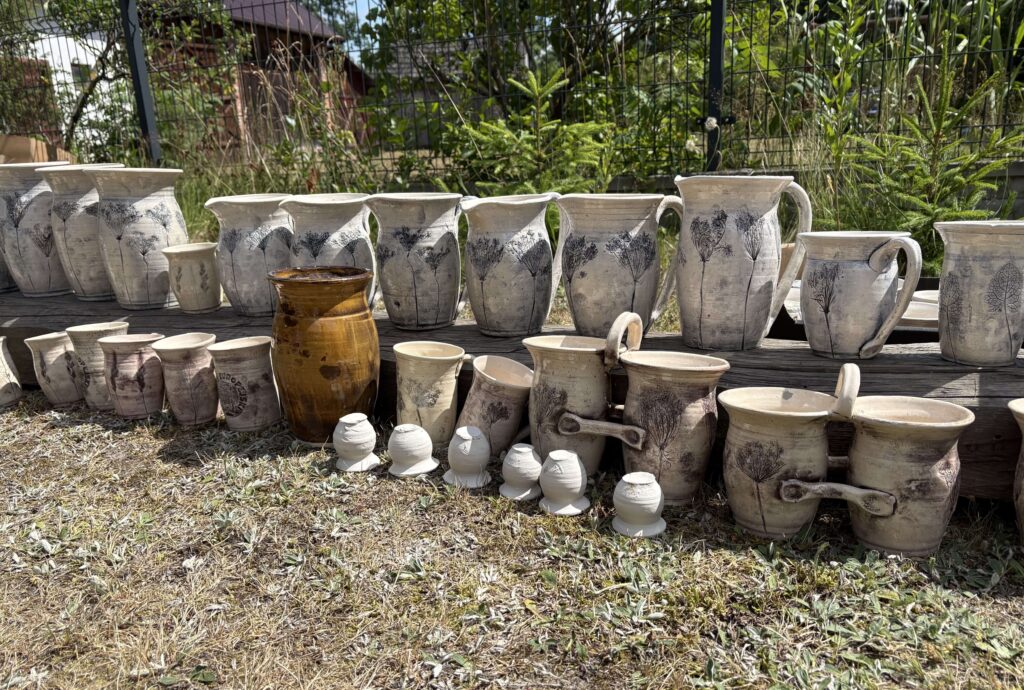 Ceramiczne wazony i dzbanki w kolorze białym i brązowym z czarnymi motywami roślinnymi ustawione na drewnianej ławce na tle zieleni i ogrodzenia.