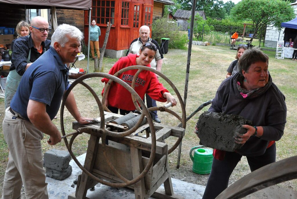 Grupa ludzi wokół drewnianej prasy, jedna osoba trzyma szarą cegłę z gliny, otoczenie z zielenią i drewnianymi budynkami.