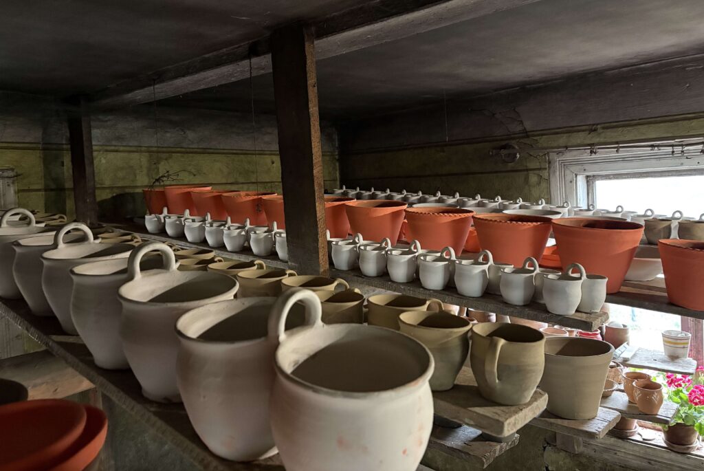 Wiele ceramicznych, nieglazurowanych garnków i doniczek w różnych rozmiarach ustawionych na drewnianych półkach w ciemnym pomieszczeniu.