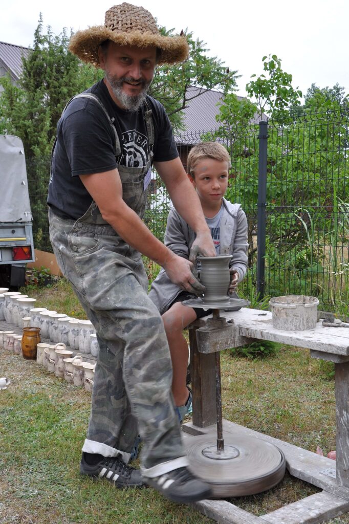 Mężczyzna w kapeluszu i chłopiec formują na kole garncarskim szarą glinianą wazę, wokół stoją gotowe naczynia, w tle zielone krzewy i metalowy płot.