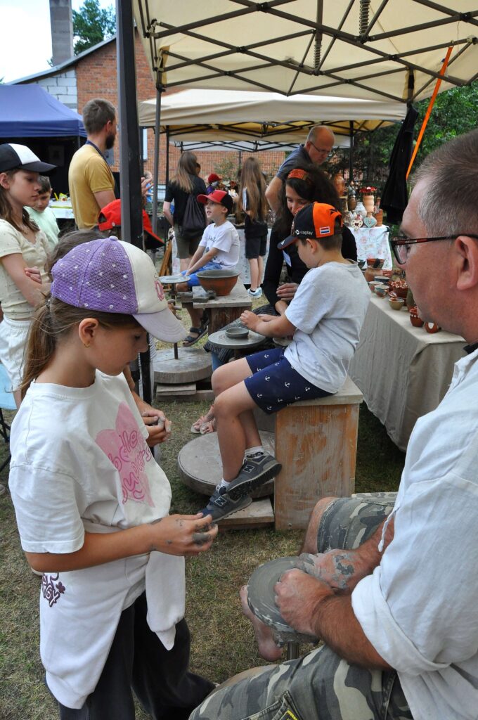 Dzieci i dorośli pod beżowymi namiotami, chłopiec i mężczyzna przy kole garncarskim, stół z ceramiką, tło z ceglanym budynkiem i zielenią.