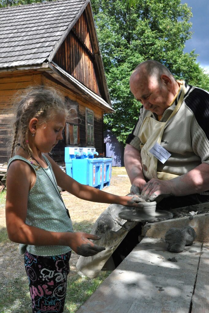 Dziewczynka z warkoczami i mężczyzna z gołą głową formujący glinę na kole garncarskim na tle drewnianego domu i zielonych drzew.