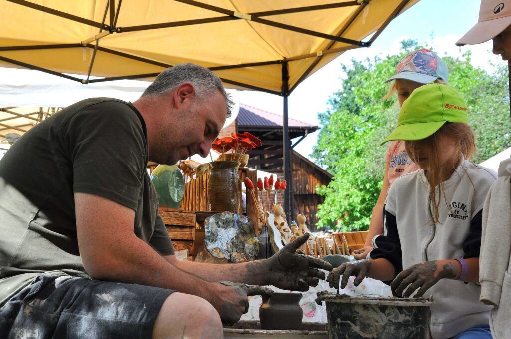 Mężczyzna i dwie dziewczynki pod żółtym namiotem formują gliniane naczynie na kole garncarskim, obok ceramiczne figurki i dekoracje, w tle zielone drzewa i drewniany budynek.