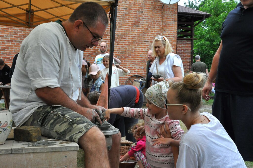 Mężczyzna formujący glinę na kole garncarskim, dziecko i kobieta obok, w tle ludzie pod namiotem i ceglana ściana budynku.