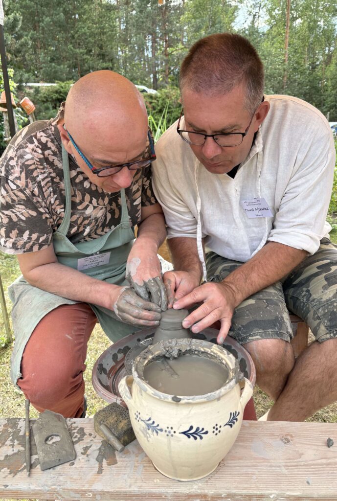 Dwóch mężczyzn z okularami formuje mokrą glinę na kole garncarskim, drewniany stół z narzędziami i ceramiczny dzbanek z wodą, w tle zielone drzewa.