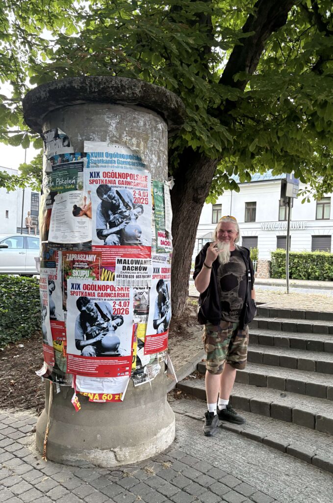 Cylindryczna tablica ogłoszeń z licznymi plakatami, w tym dużymi dotyczącymi spotkań garncarskich, obok stoi mężczyzna w czarnej kamizelce, moro spodniach i czarnych butach, za nim schody i drzewa.