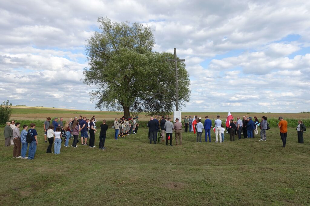 Grupa ludzi stojąca na trawie wokół dużego drzewa i drewnianego krzyża na tle pola i nieba z chmurami. Niektórzy trzymają flagi Polski.