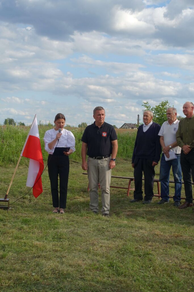 Pięć osób stojących na trawie, kobieta z mikrofonem obok czerwono-biało-czerwonej flagi, mężczyźni w różnokolorowych ubraniach, zielony teren i niebo z chmurami w tle.