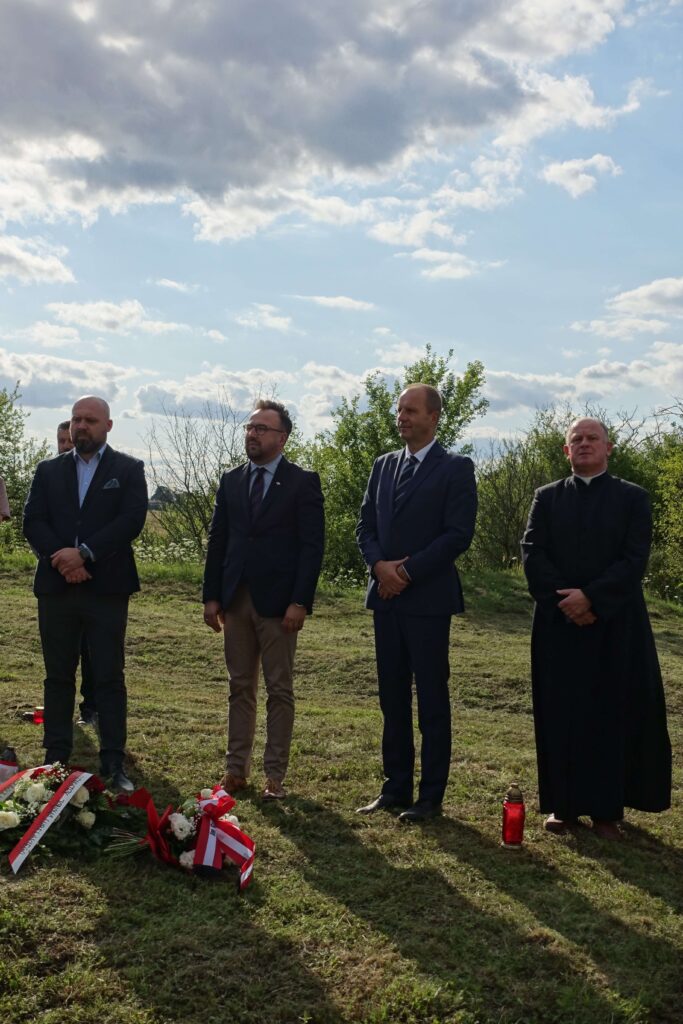 Czterech mężczyzn w garniturach i sutannie stoi na trawie, przed nimi wiązanki kwiatów i znicze, w tle drzewa i niebieskie niebo z chmurami.
