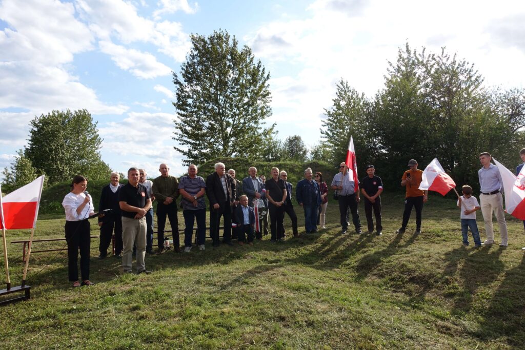 Grupa ludzi stojących na trawie na tle drzew, kilka osób trzyma flagi Polski, kobieta mówi do mikrofonu na stojaku.