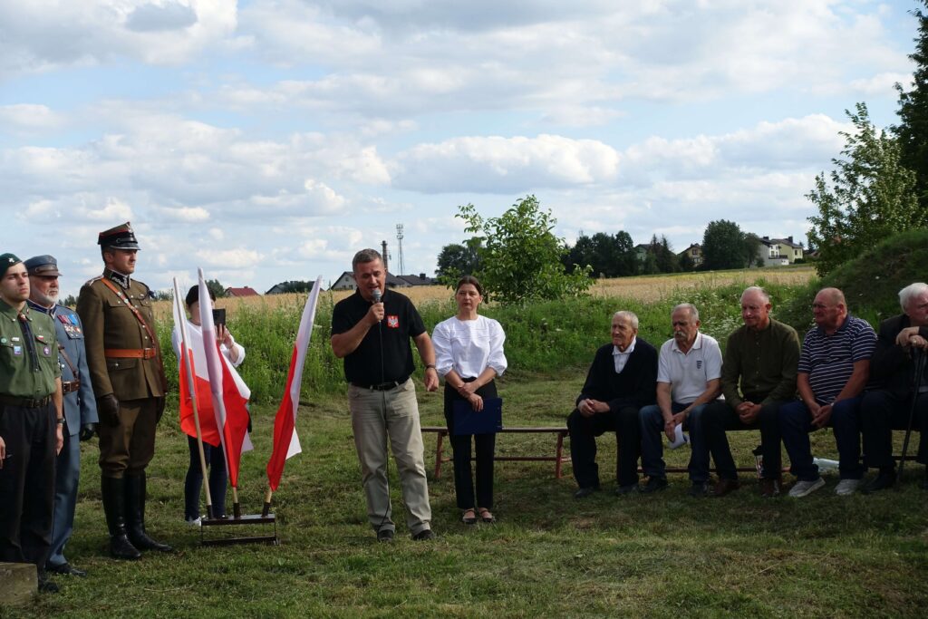 Grupa ludzi na zewnątrz, część w mundurach wojskowych, mężczyzna przemawia do mikrofonu, biało-czerwone flagi, zielone pole i niebieskie niebo z chmurami.