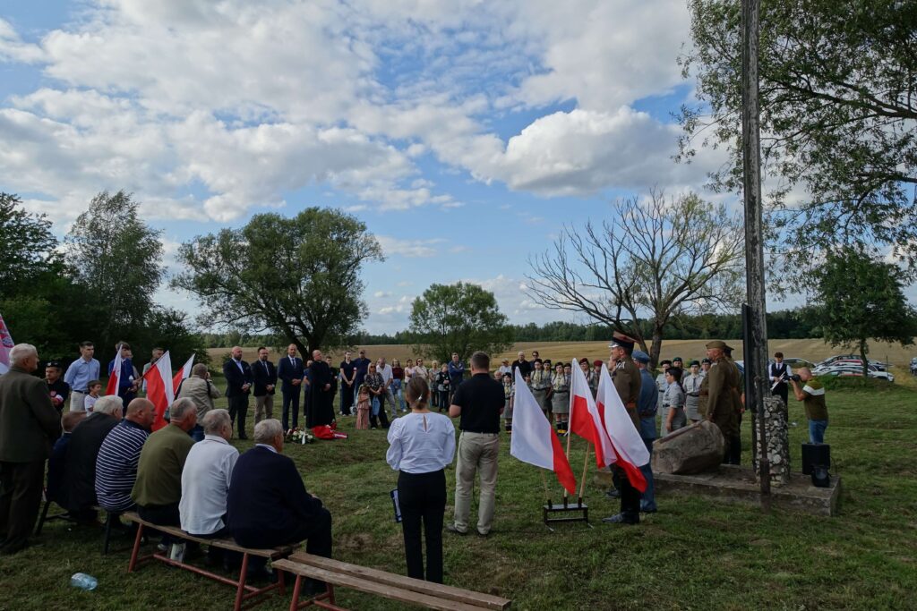 Ludzie stojący i siedzący na trawie i ławkach, kilka osób w mundurach, biało-czerwone flagi, drzewa i pola, niebieskie niebo z chmurami.