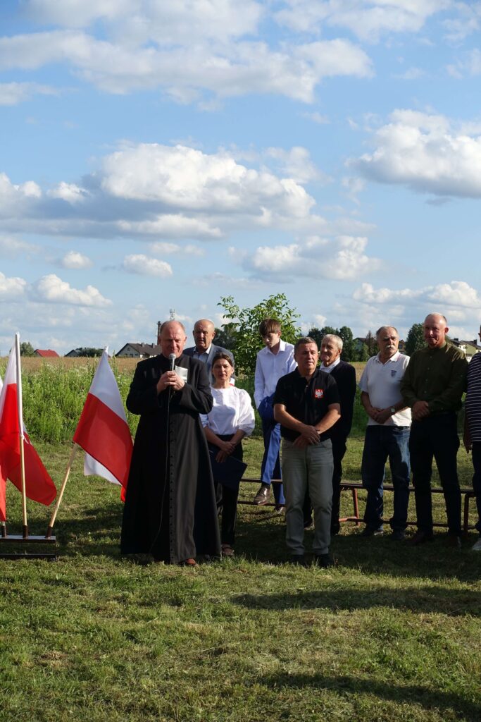 Grupa ludzi na zewnątrz na tle zielonej łąki i nieba z chmurami, mężczyzna w sutannie z mikrofonem, dwie flagi Polski.