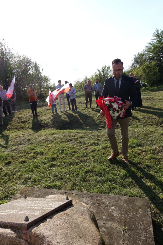 Mężczyzna w ciemnej marynarce i okularach trzyma bukiet kwiatów z biało-czerwonymi wstążkami, grupa ludzi z flagami Polski w tle, zielona trawa, kamienna płyta pomnika.