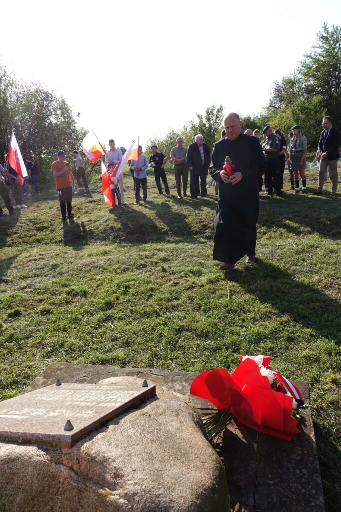 Grupa ludzi stojących na trawie z flagami Polski, mężczyzna w sutannie idzie z czerwonym zniczem, na pierwszym planie kamień z tablicą i bukiet biało-czerwonych kwiatów.