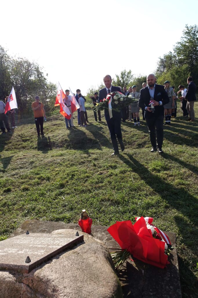 Grupa ludzi na trawie z biało-czerwonymi flagami, mężczyźni w garniturach niosący wiązanki kwiatów, w tle drzewa, na pierwszym planie kamień z tablicą i czerwona lampka.