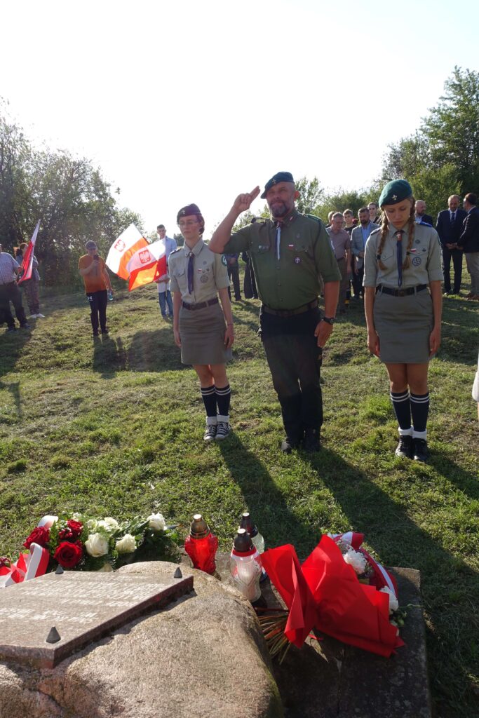 Trzy osoby w mundurach harcerskich, dwie kobiety i mężczyzna, stoją na trawie przy kamieniu pamiątkowym z kwiatami i zniczami, w tle ludzie i flagi Polski.