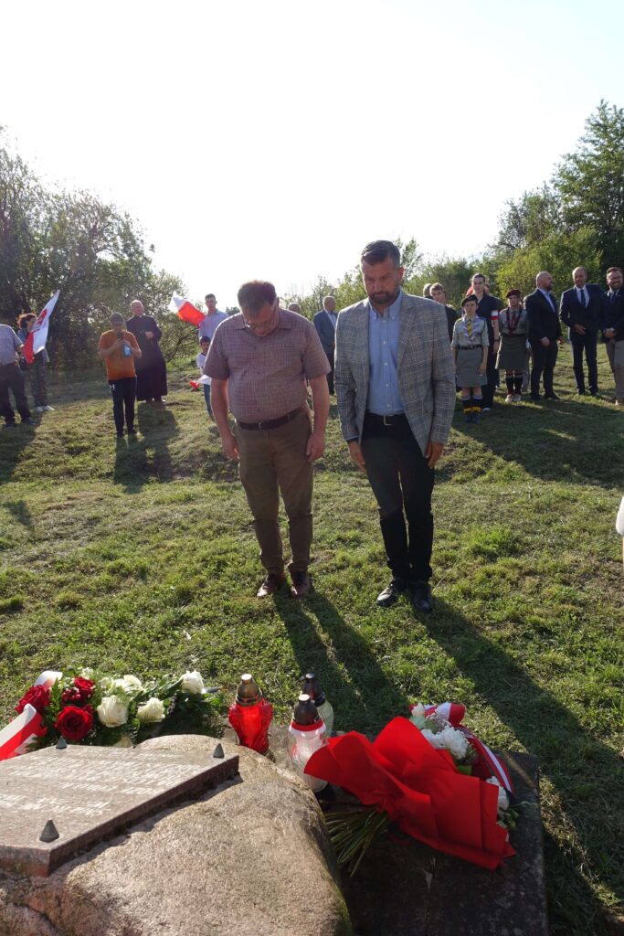 Dwóch mężczyzn opuszcza głowy przed kamieniem z tablicą pamiątkową, wokół grupa ludzi, kwiaty czerwono-białe i znicze na trawie.