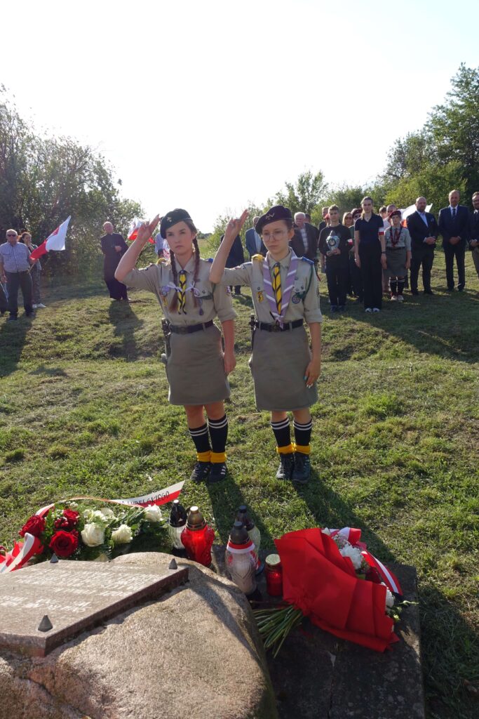 Dwie harcerki w beżowych mundurach i beretach, stoją na trawie przy kamieniu pamiątkowym z kwiatami i zniczami, w tle grupa ludzi na tle drzew i nieba.