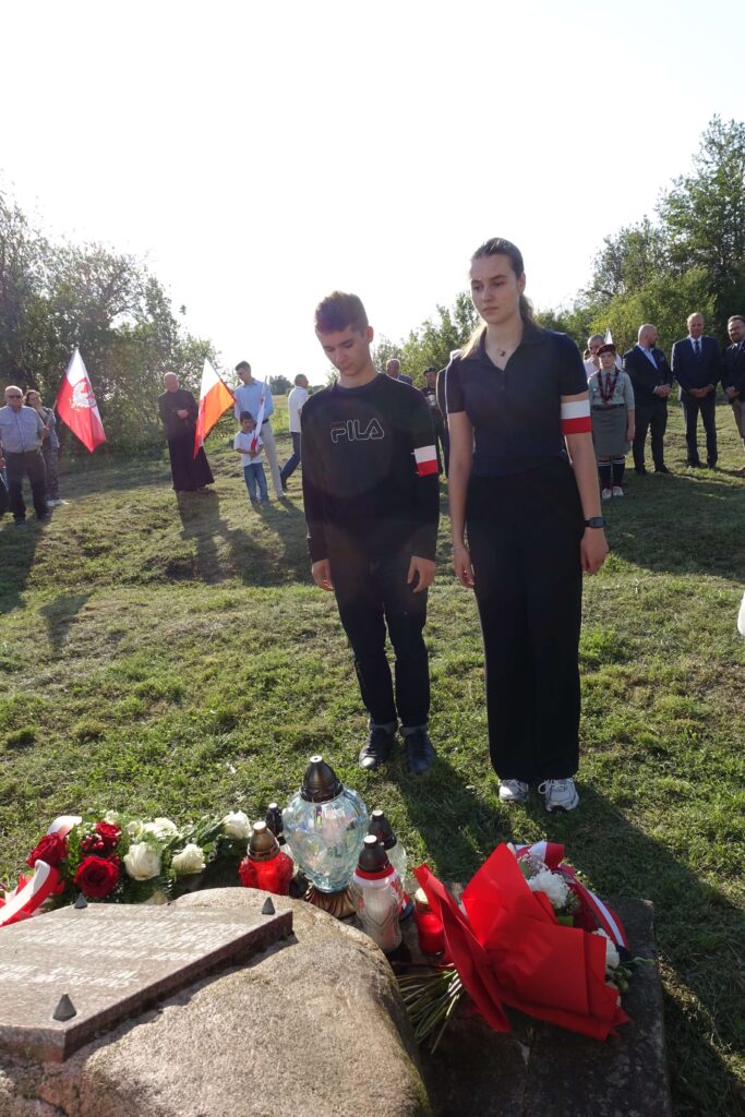 Młody chłopak i dziewczyna z opaskami biało-czerwonymi stojący na trawie przy kamieniu z tablicą, przed nimi kwiaty i znicze, w tle ludzie z flagami Polski i zieleń.