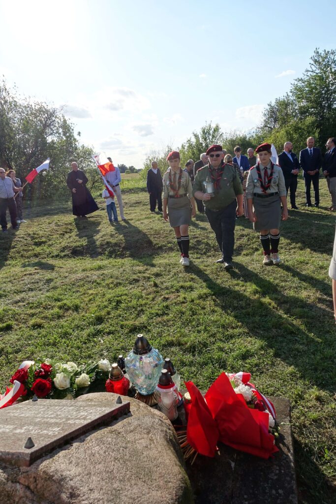 Grupa ludzi na trawie, w centrum mężczyzna i dwie dziewczyny w mundurach harcerskich z czerwonymi beretami, z przodu kamień z lampionami i kwiatami, w tle drzewa i niebieskie niebo.